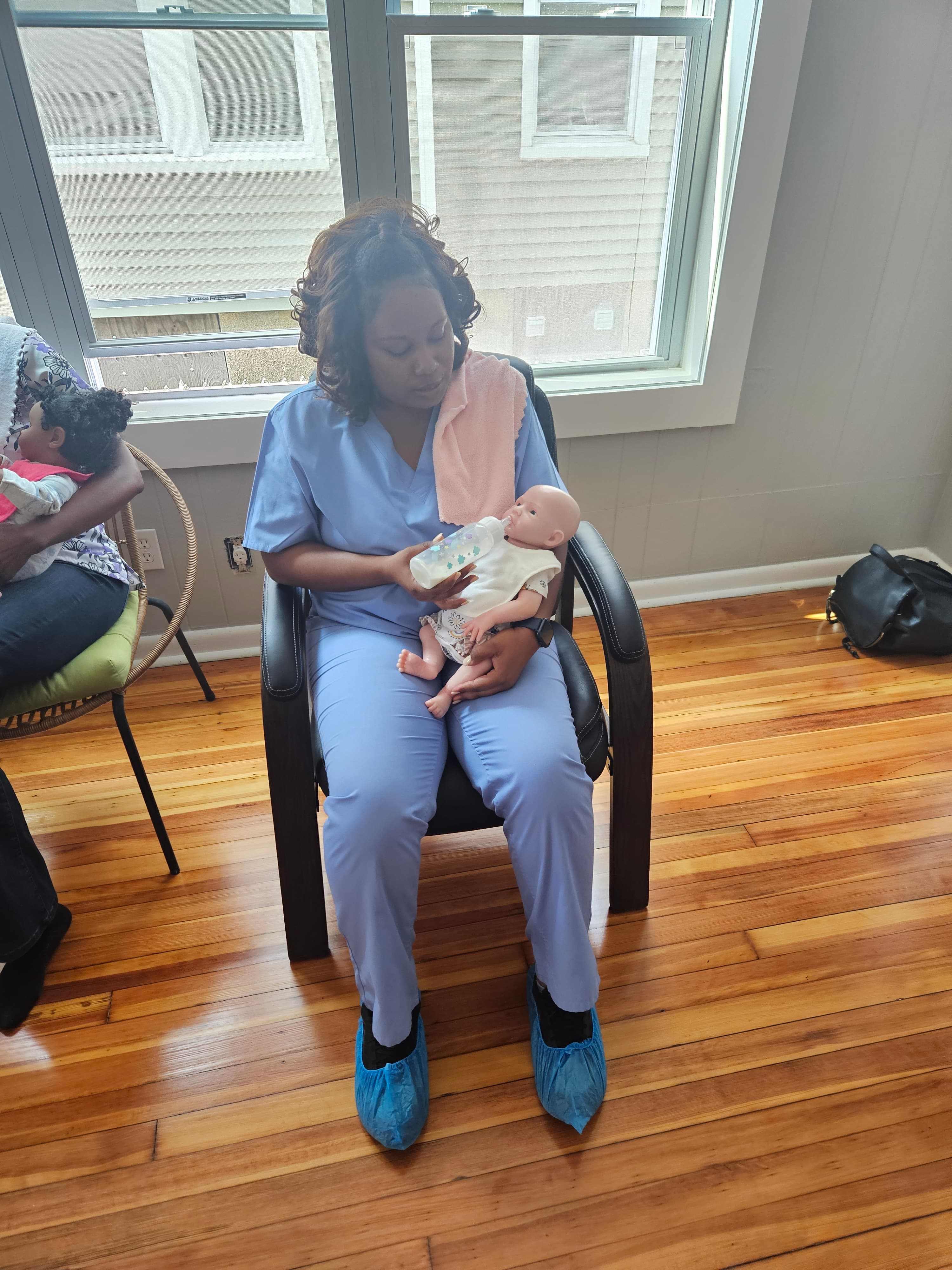 Woman in blue scrubs sits in a chair, bottle-feeding a lifelike infant training mannequin.